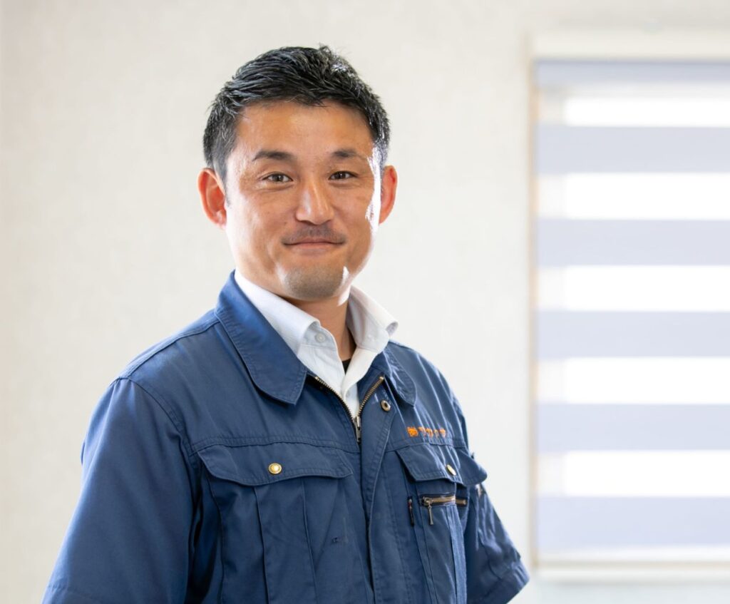 写真：代表取締役　安本昌広（やすもとまさひろ）　作業着に身を包み、笑顔でこちらを見つめている。