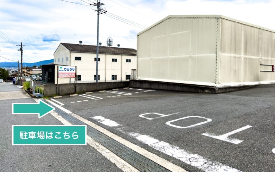 駐車場の位置を示す写真。窓のないクリーム色の壁の建物の横に、「ワカクサ」の文字が書かれた看板が立っており、その前に車が3台停められるようになっている。